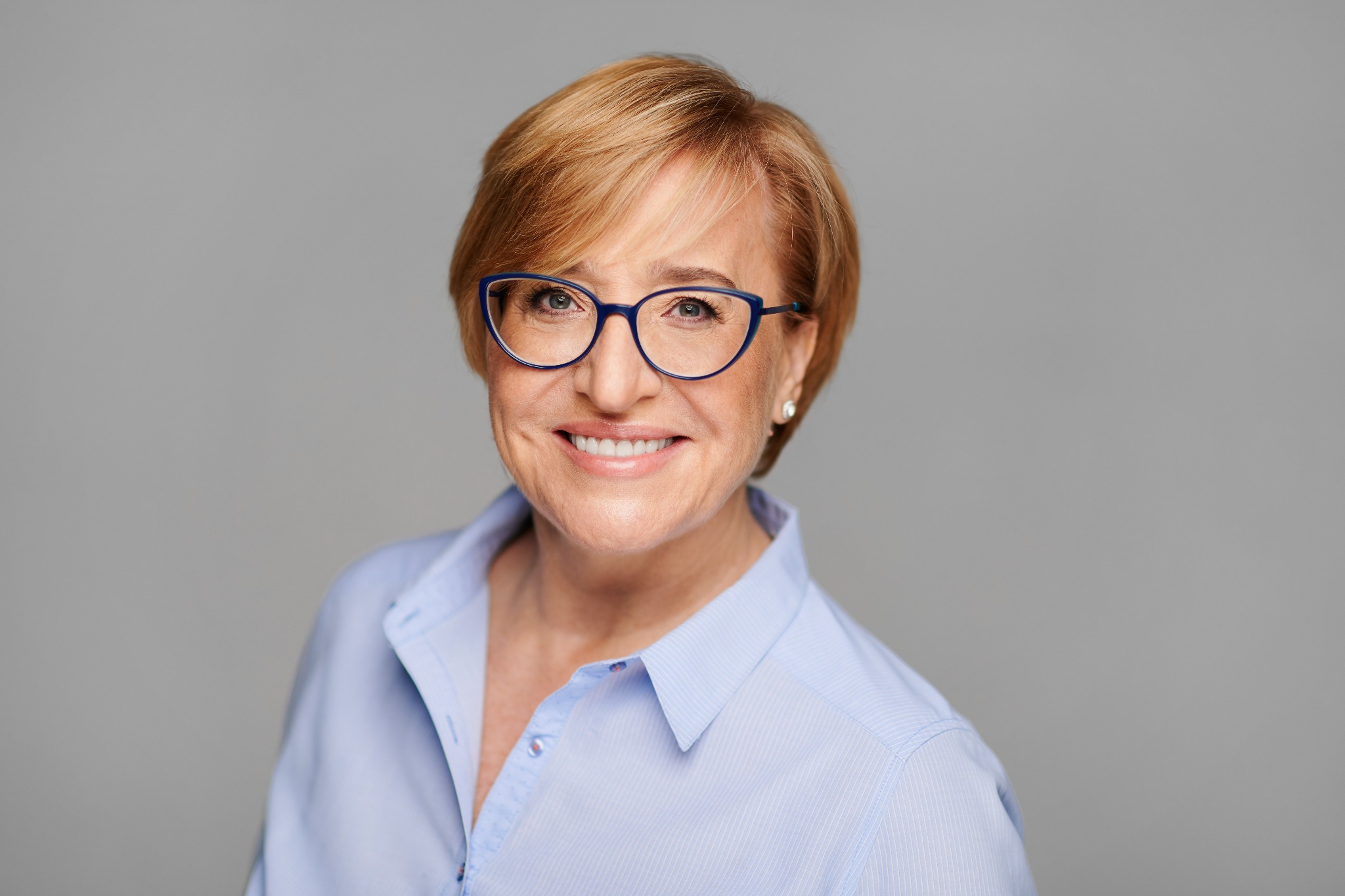 Portret uśmiechniętej kobiety w okularach, z krótkimi blond włosami i błękitnej koszuli na jasnoszarym tle.