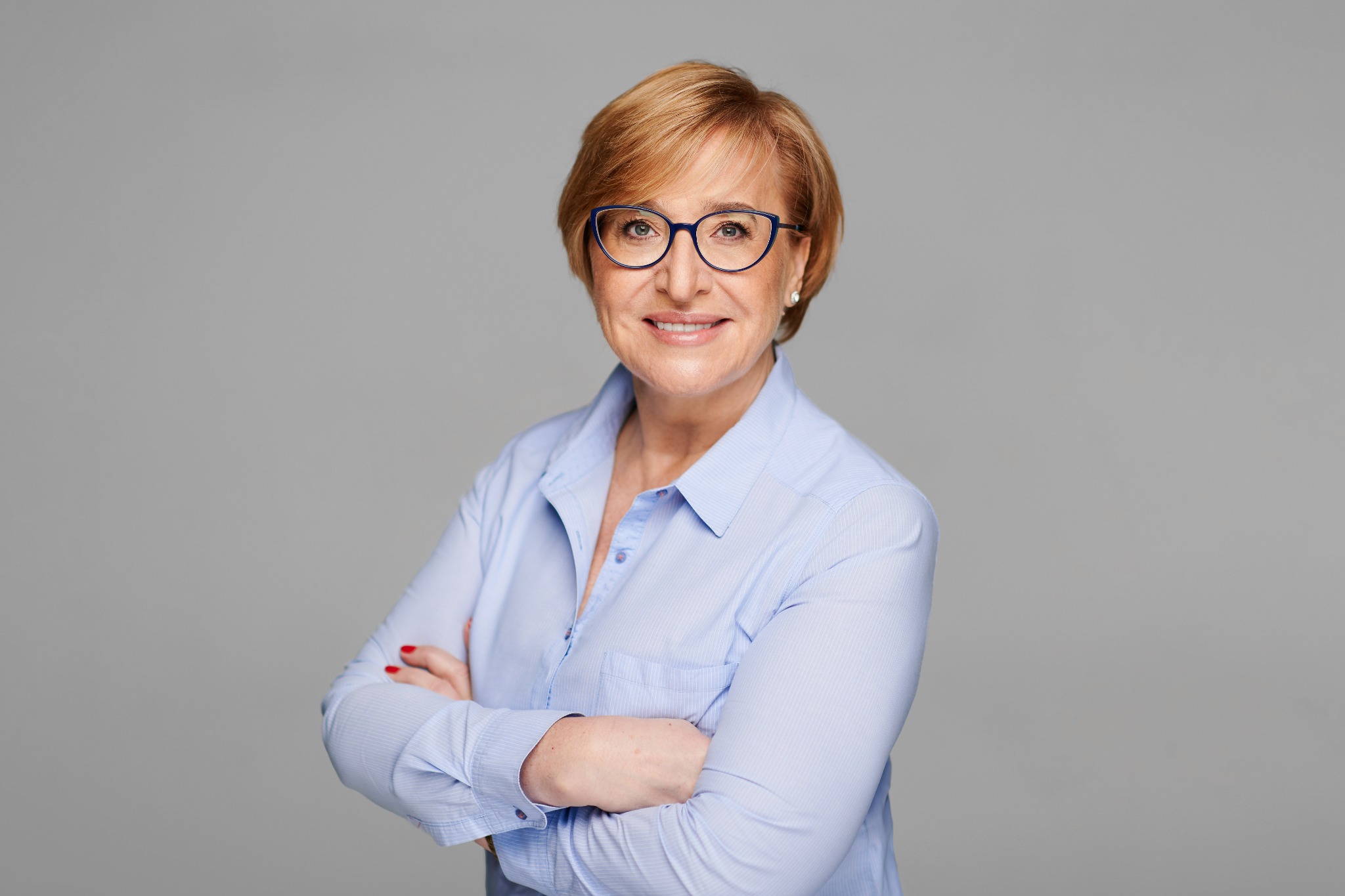 Portret uśmiechniętej kobiety w okularach i błękitnej koszuli, z założonymi rękami na szarym tle.