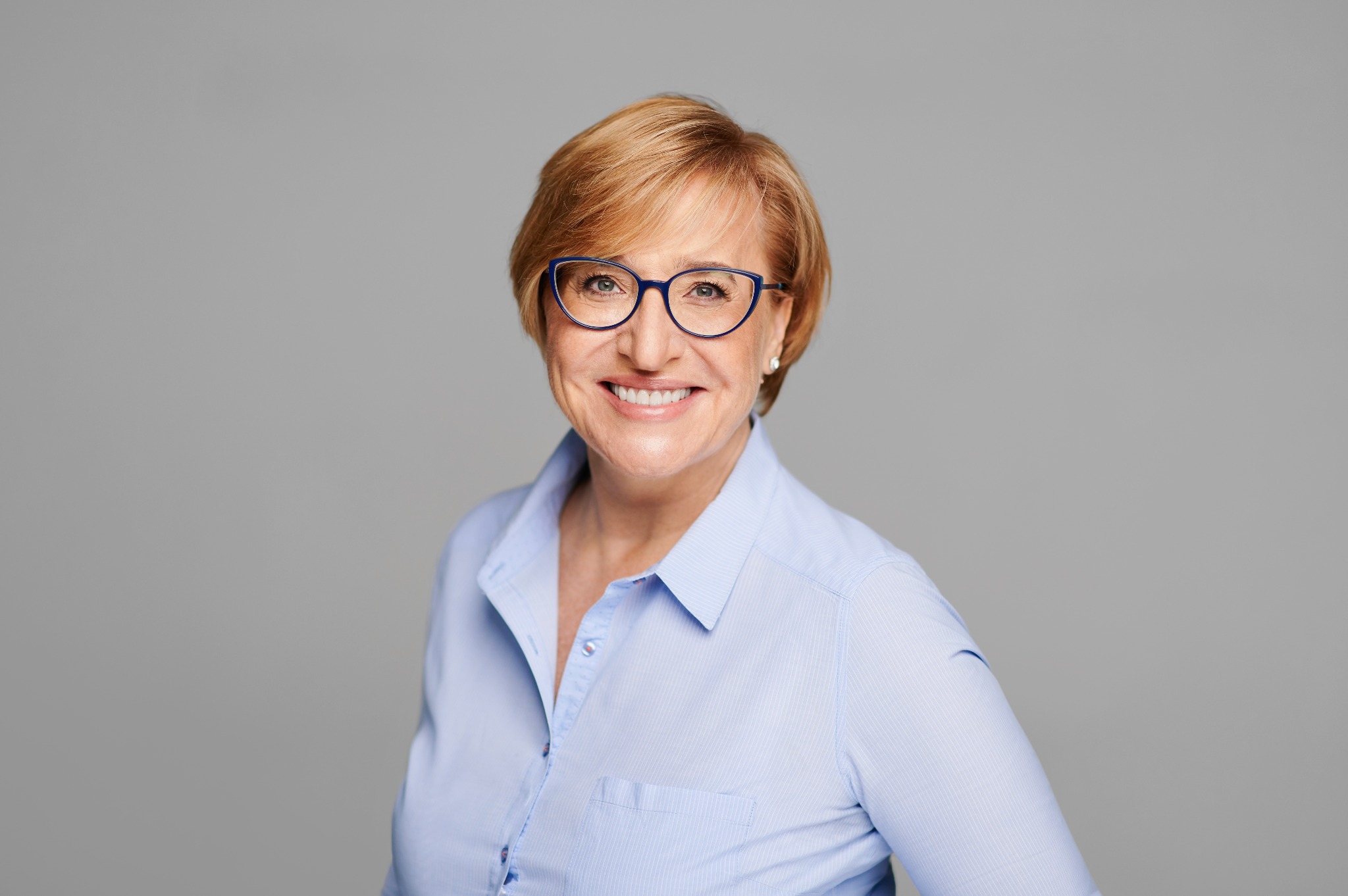 Portret uśmiechniętej kobiety w okularach z niebieskimi oprawkami i błękitnej koszuli na jednolitym szarym tle.