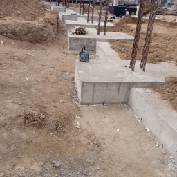 Wykopane fundamenty z wystającymi prętami zbrojeniowymi i poziomnicą laserową Makita na betonowym fundamencie, na placu budowy z koparką w tle.