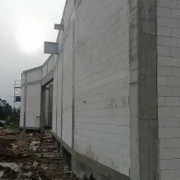 Wznoszenie ściany budynku z białych bloczków, z widocznymi żelbetowymi słupami wzmacniającymi, na etapie surowym otwartym, z widocznym gruntem wokół fundamentów.