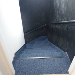 Wojtek - Klatka schodowa z niebieską wykładziną dywanową, ciemnoszara ściana z efektem dekoracyjnym, biała ściana boczna i drewniana podłoga u dołu schodów.