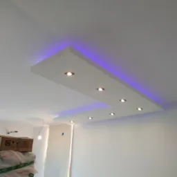 Podwieszany sufit z oświetleniem punktowym i dekoracyjnym niebieskim podświetleniem LED, widoczne materiały budowlane w rogu pomieszczenia.