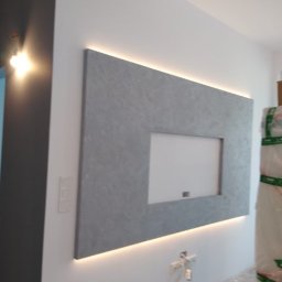 Wojtek - Szara, dekoracyjna ściana z podświetleniem LED i wnęką na telewizor, widoczne gniazdka elektryczne i materiały budowlane w folii.