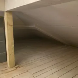 Poddasze w trakcie prac wykończeniowych: widoczne płyty kartonowo-gipsowe na skosach dachu, drewniana konstrukcja wsporcza i surowa drewniana podłoga z desek.