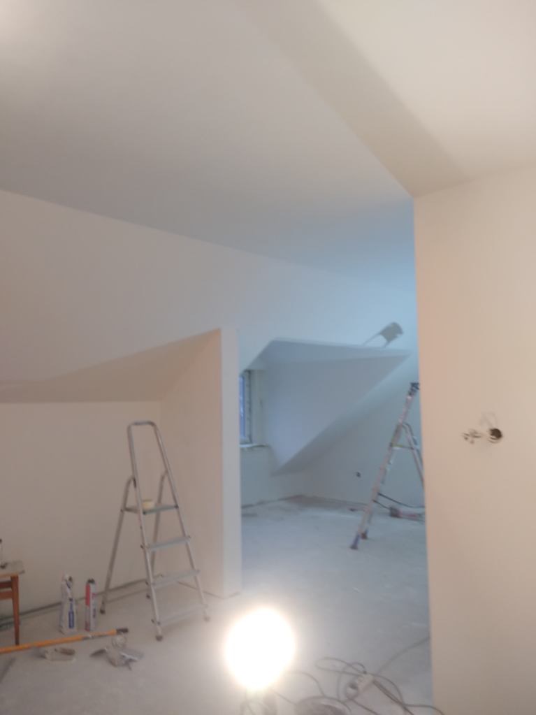 Poddasze po remoncie: świeżo pomalowane ściany i sufit na biało, dwie aluminiowe drabiny, widoczne narzędzia malarskie i lampa oświetlająca podłogę.