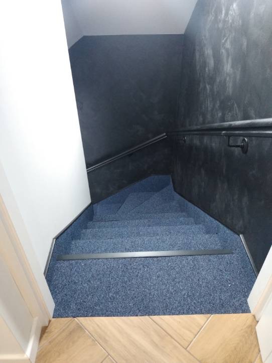 Klatka schodowa z niebieską wykładziną dywanową, ciemnoszara ściana z efektem dekoracyjnym, biała ściana boczna i drewniana podłoga u dołu schodów.