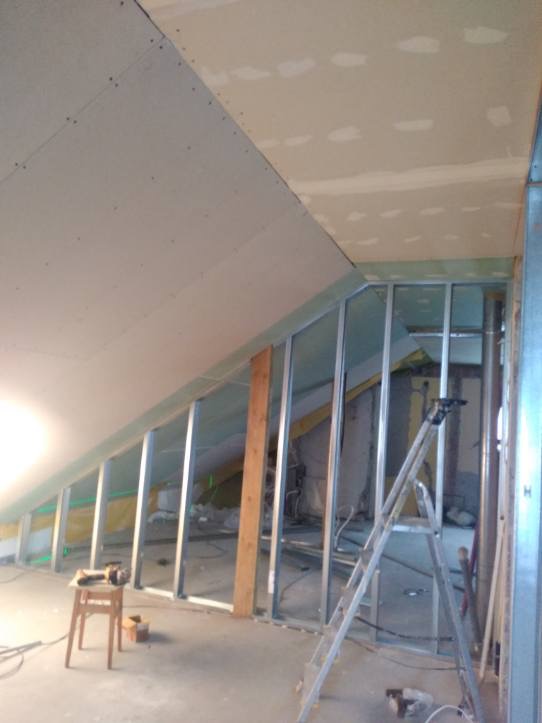 Instalacja płyty karton gips na poddaszu ze skośnym sufitem, widoczna metalowa konstrukcja, drabina i narzędzia na stołku.