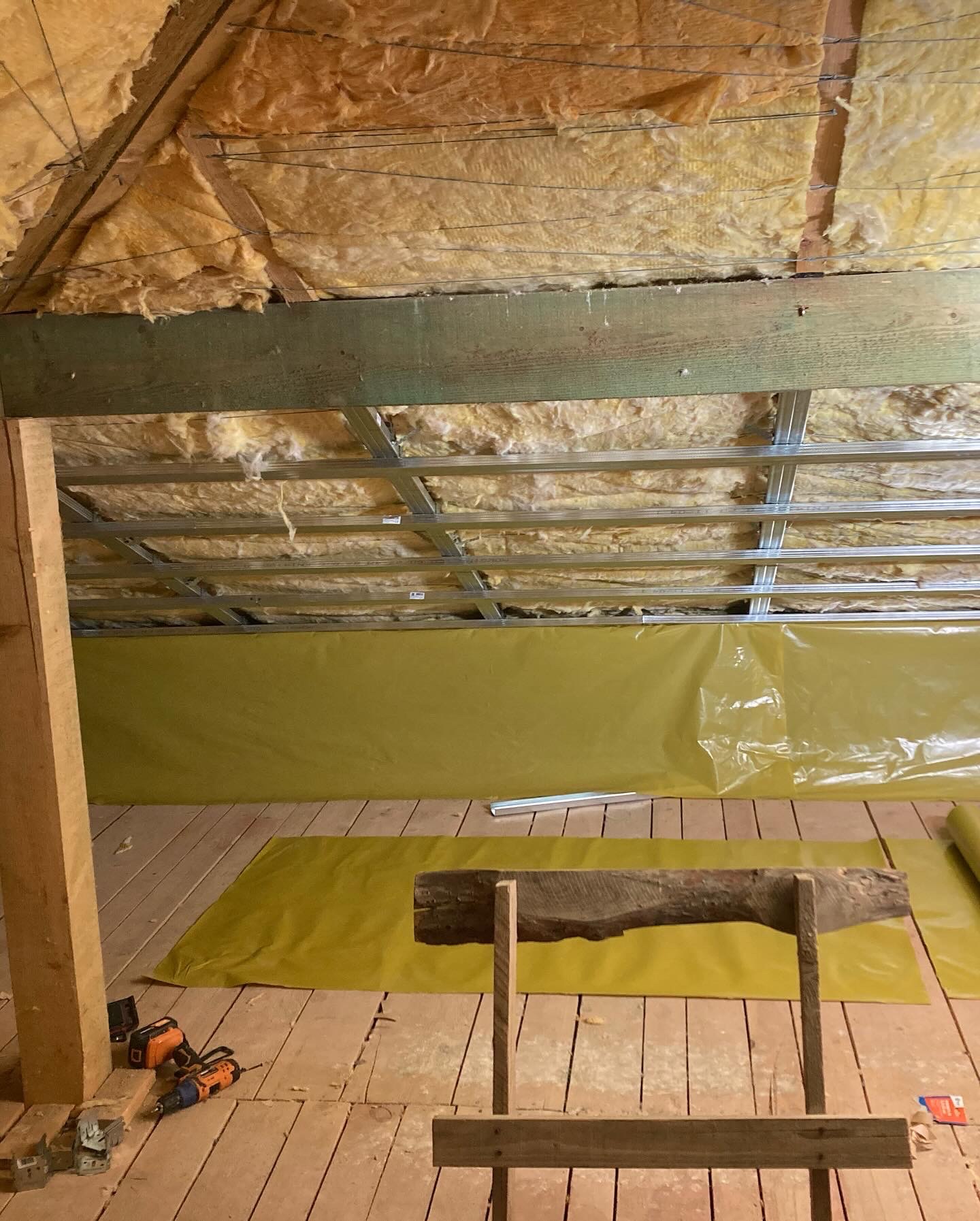 Poddasze w trakcie ocieplania: wełna mineralna na skosach dachu, metalowy stelaż pod płytami, żółta folia paroizolacyjna na ścianach, drewniana podłoga, narzędzia i prowizoryczny stojak