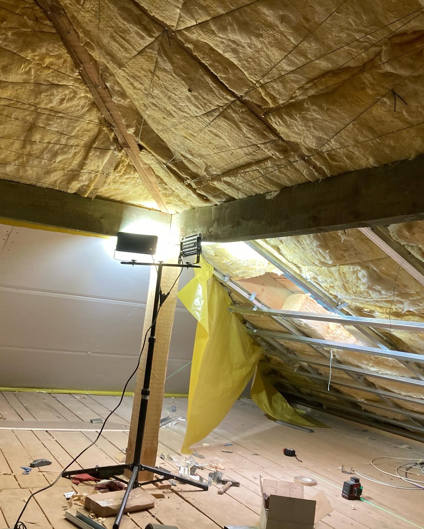 Poddasze w trakcie ocieplania wełną mineralną, widoczne metalowe profile konstrukcyjne, reflektor na statywie oświetla przestrzeń, na podłodze narzędzia i resztki materiałów budowlanych.
