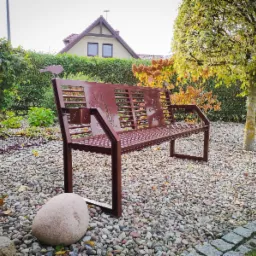 Metalowa ławka ogrodowa w kolorze brązowym, z ażurowym oparciem z motywami roślinnymi i ptakiem na szczycie, na tle ogrodu z kamieniami i krzewami.