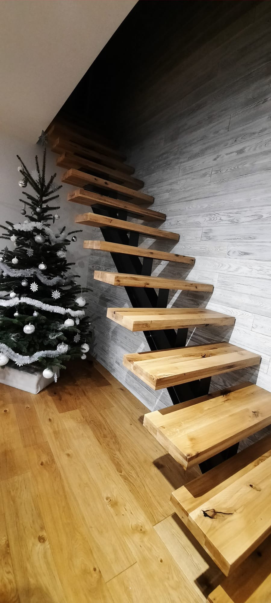 Drewniane schody wspornikowe z czarną metalową konstrukcją, obok choinka z srebrnymi ozdobami, na tle ściany z paneli imitujących drewno.