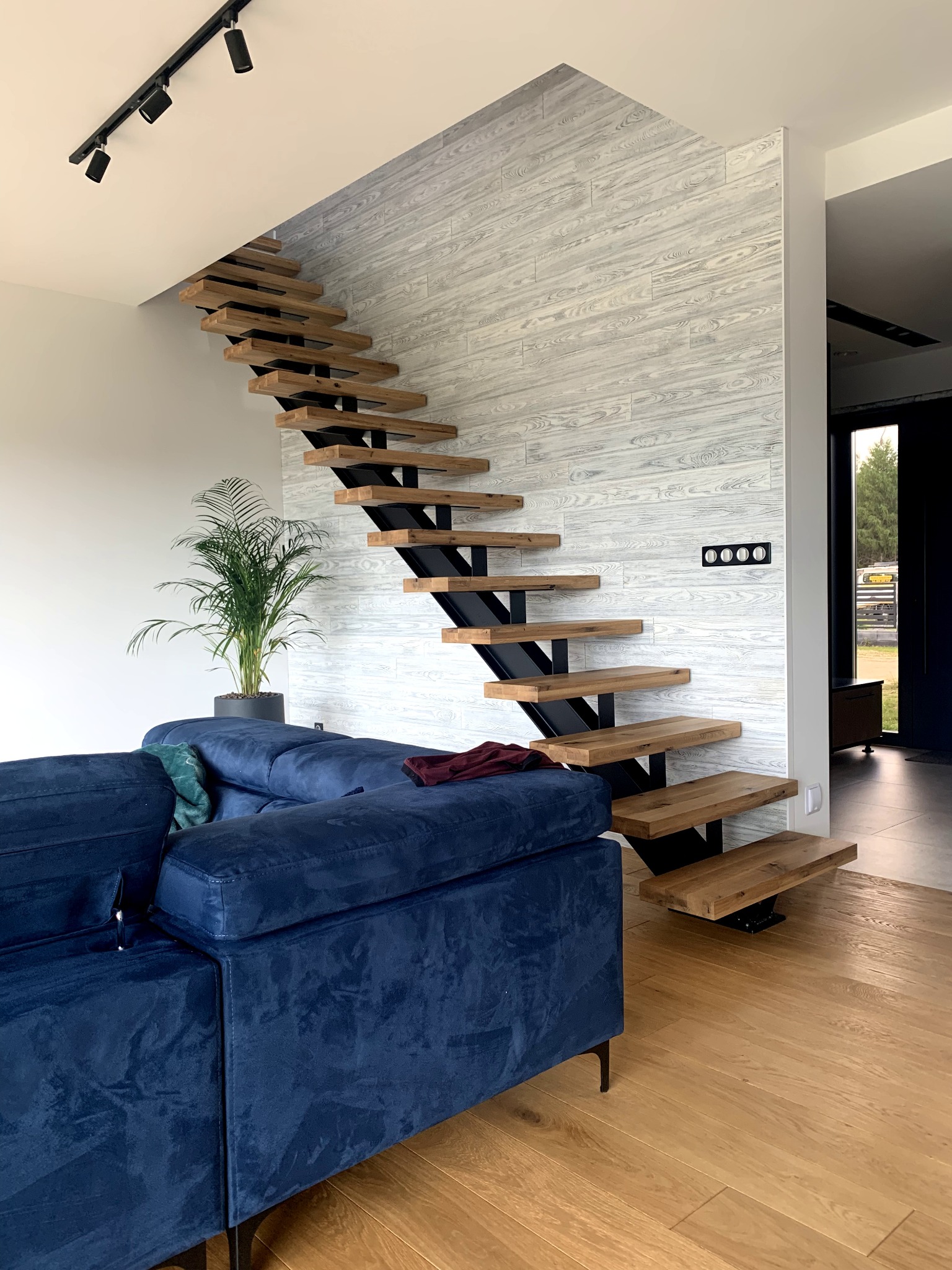 Nowoczesne schody z drewnianymi stopniami i czarną metalową konstrukcją w jasnym wnętrzu z drewnianą podłogą i niebieską sofą.
