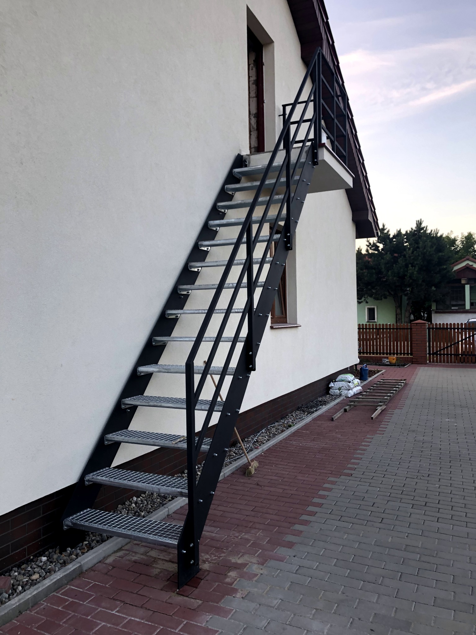 Zewnętrzne schody metalowe z kratowymi stopniami, prowadzące do drzwi na piętrze budynku z białymi ścianami, widoczne worki z zaprawą i drabina oparta o ścianę.