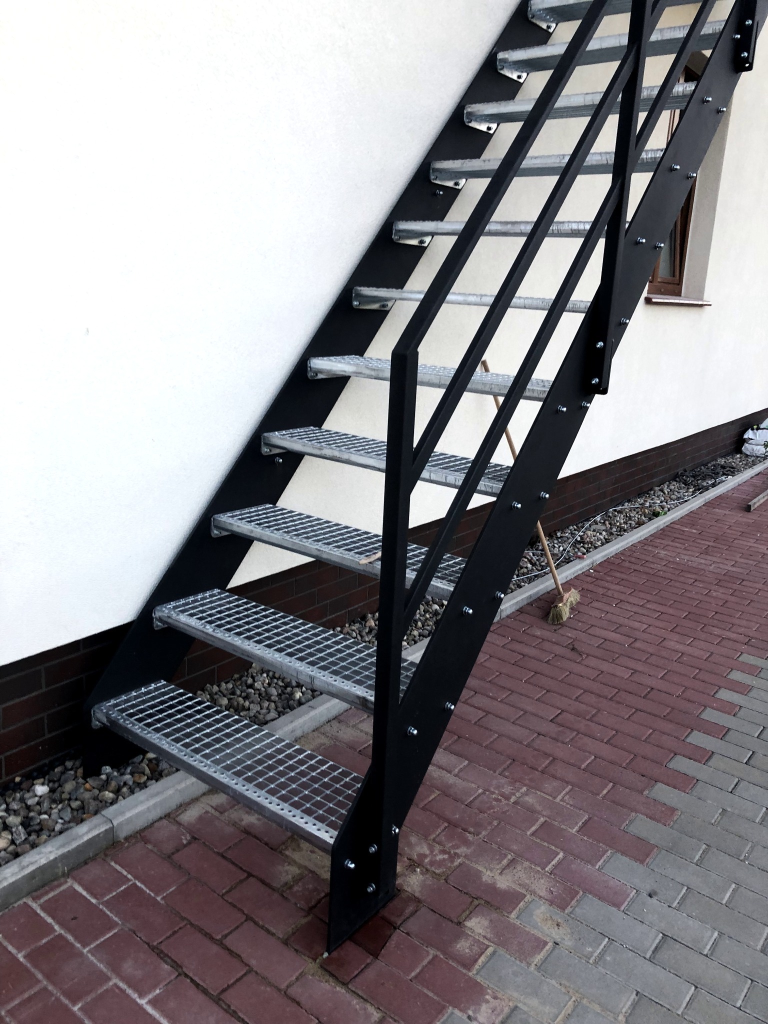 Zewnętrzne schody stalowe z kratowanymi stopniami, mocowane do białej ściany budynku, z widoczną kostką brukową na podłożu i miotłą opartą o konstrukcję.