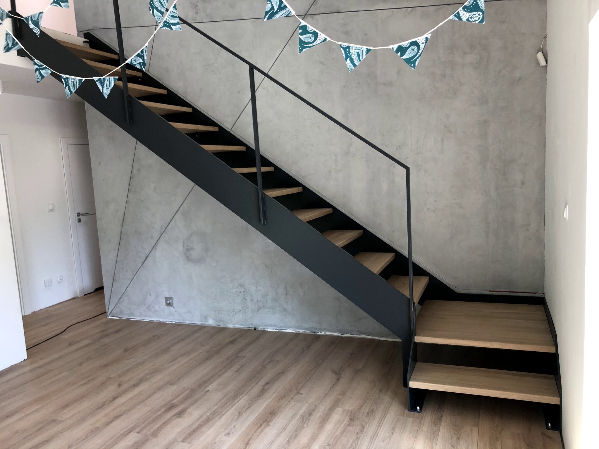 Nowoczesne schody z drewnianymi stopniami i czarną metalową konstrukcją, wiszące dekoracje z trójkątnych chorągiewek, surowa ściana z efektem betonu.