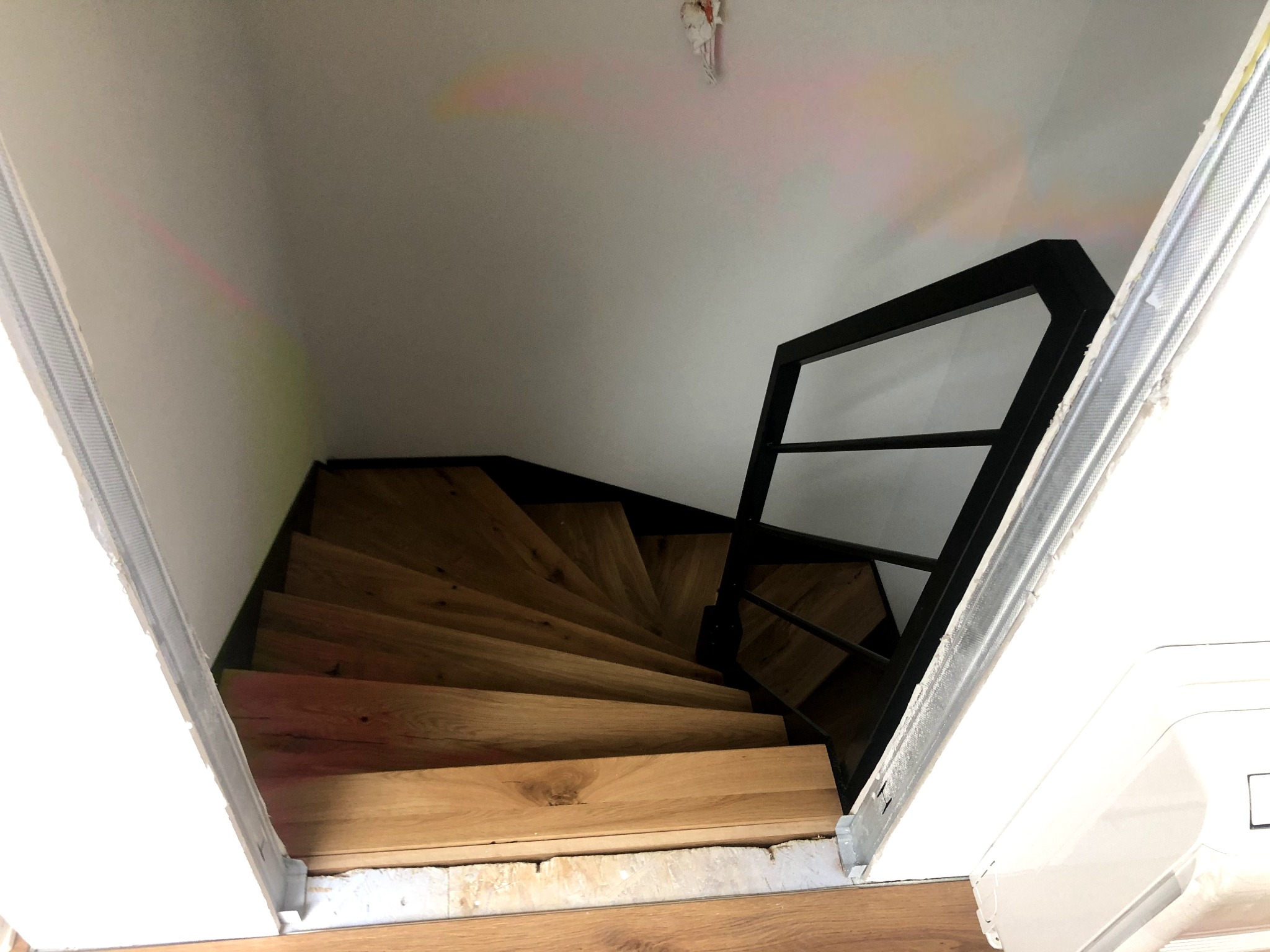 Widok z góry na drewniane schody spiralne z czarną, metalową balustradą, w trakcie wykańczania, widoczne surowe krawędzie ścian i podłogi.