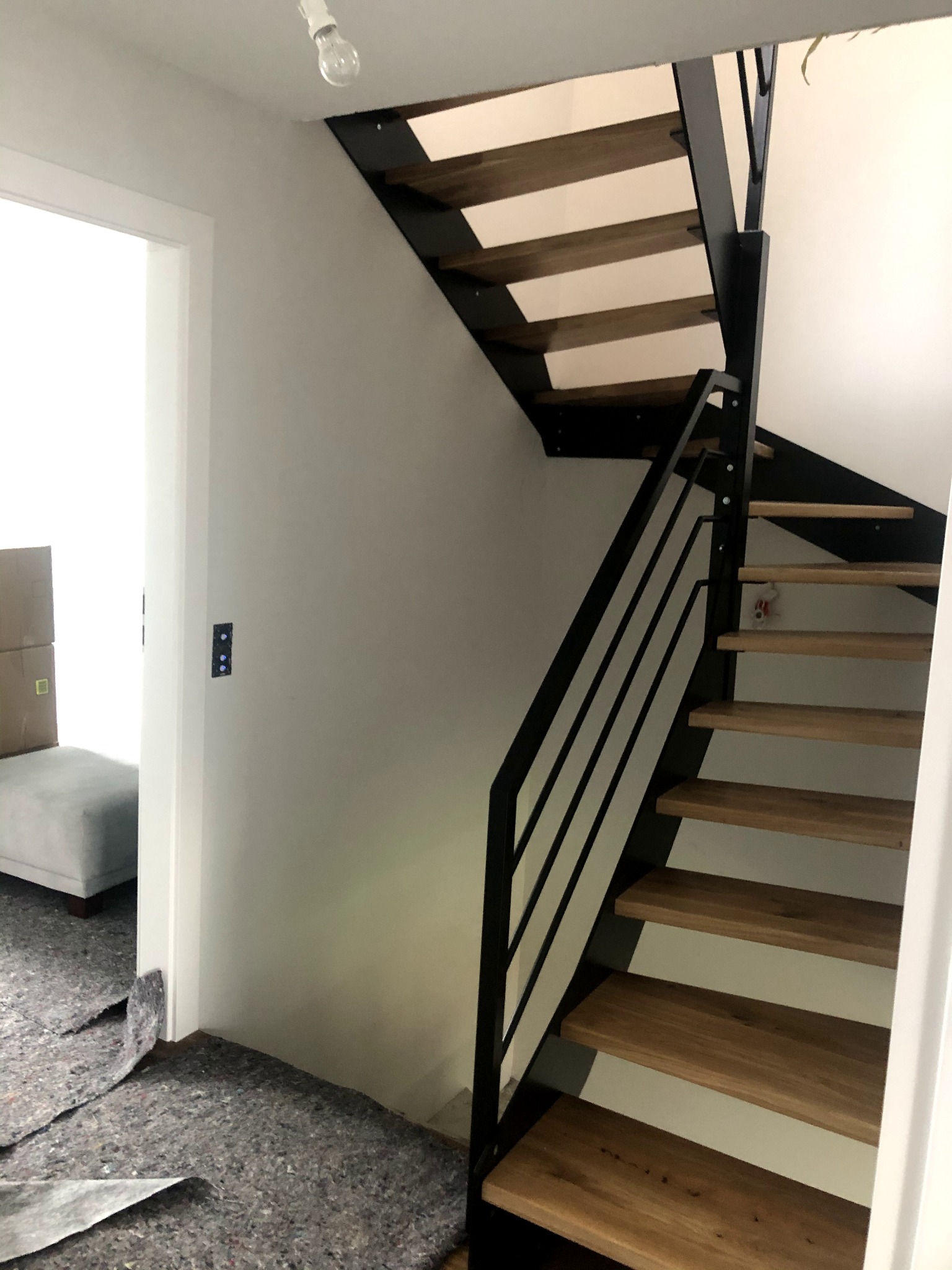 Nowoczesne schody z drewnianymi stopniami i czarną metalową konstrukcją, widoczne z boku, z zabezpieczeniem podłogi w trakcie montażu.