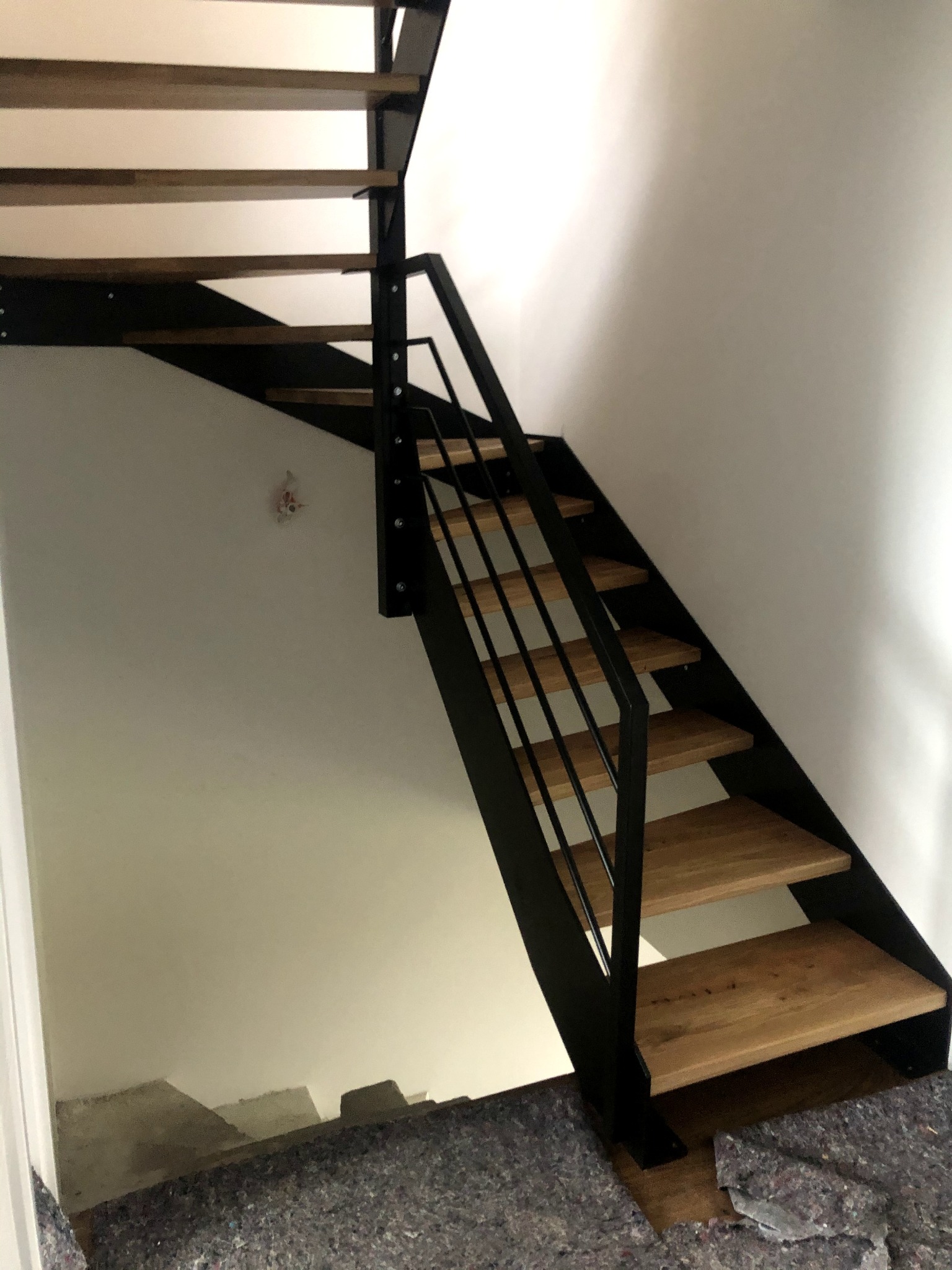 Nowoczesne schody z drewnianymi stopniami i czarną, metalową konstrukcją, widok z dołu na tle białej ściany z lekkim zabrudzeniem.