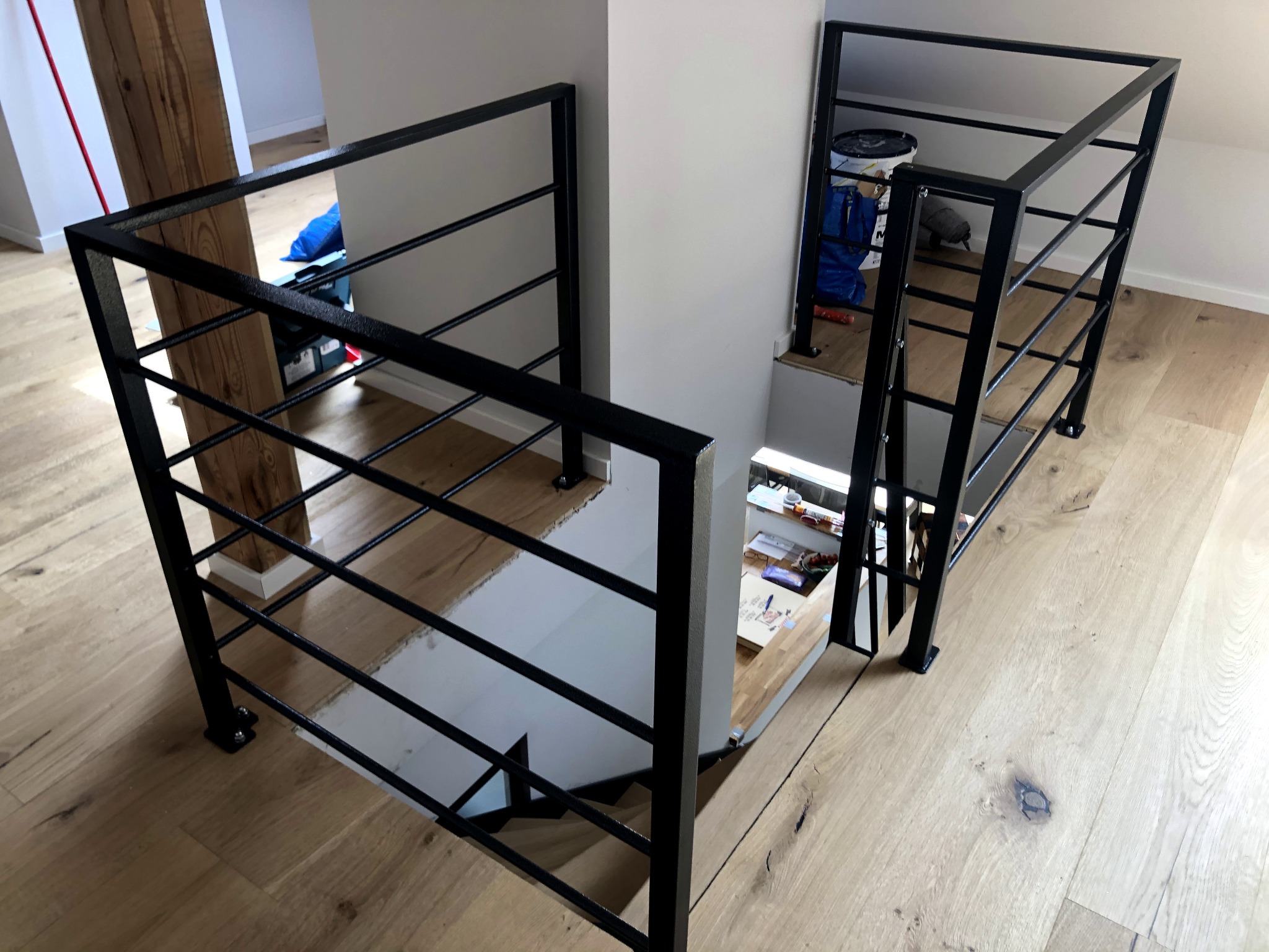 Nowoczesna, czarna balustrada schodowa z metalowymi prętami, widok z góry na drewniane schody i podłogę, z elementami wyposażenia wnętrza w tle.