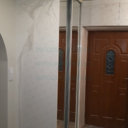 Infoduo Spółka z Ograniczoną Odpowiedzialnością - Nowa biała szafa z przesuwnymi lustrzanymi drzwiami, częściowo zabezpieczona folią ochronną, zamontowana we wnęce obok brązowych drzwi wejściowych.