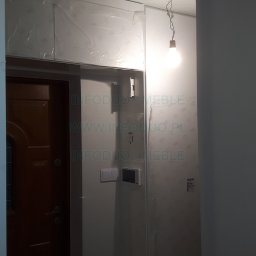 Infoduo Spółka z Ograniczoną Odpowiedzialnością - Montaż szafek pod sufitem w Lubartowie, zabezpieczonych folią ochronną, widoczny również fragment brązowych drzwi i surowa żarówka na kablu.