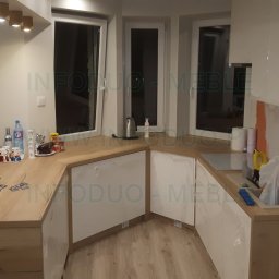 Infoduo Spółka z Ograniczoną Odpowiedzialnością - Nowoczesna kuchnia w trakcie montażu: białe szafki z drewnianymi blatami, folia ochronna na frontach, widoczne akcesoria kuchenne i okna w tle.