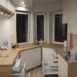 Infoduo Spółka z Ograniczoną Odpowiedzialnością - Jasna kuchnia z drewnianym blatem i białymi frontami, otwarte szuflady z systemem Blum, widoczne okna i akcesoria kuchenne na blacie.