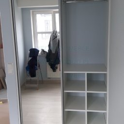 Infoduo Spółka z Ograniczoną Odpowiedzialnością - Biała szafa wnękowa z przesuwnymi drzwiami z lustrem, widoczna pusta przestrzeń z półkami i drążkiem, w lustrze odbicie pokoju z drabiną i ubraniami.