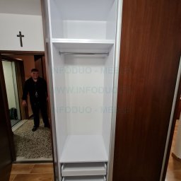 Infoduo Spółka z Ograniczoną Odpowiedzialnością - Wnętrze szafy przesuwnej z szufladami i drążkiem, widok z boku, w tle widoczna sylwetka osoby w drzwiach.