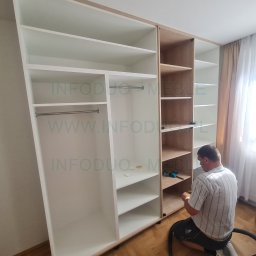 Infoduo Spółka z Ograniczoną Odpowiedzialnością - Montaż dużej, częściowo zabudowanej szafy z półkami i drążkami na ubrania, widoczny mężczyzna w trakcie pracy, narzędzia i odkurzacz na podłodze.