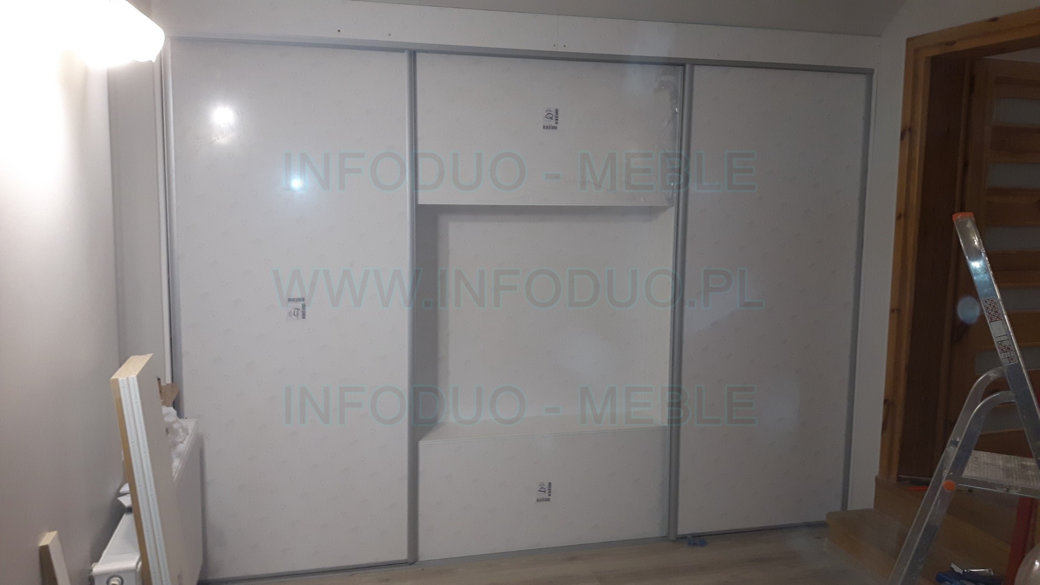 Biała szafa przesuwna na wymiar z półką w środkowej części, widoczna drabina i elementy montażowe w tle, częściowo zasłonięte materiały budowlane przy ścianie.