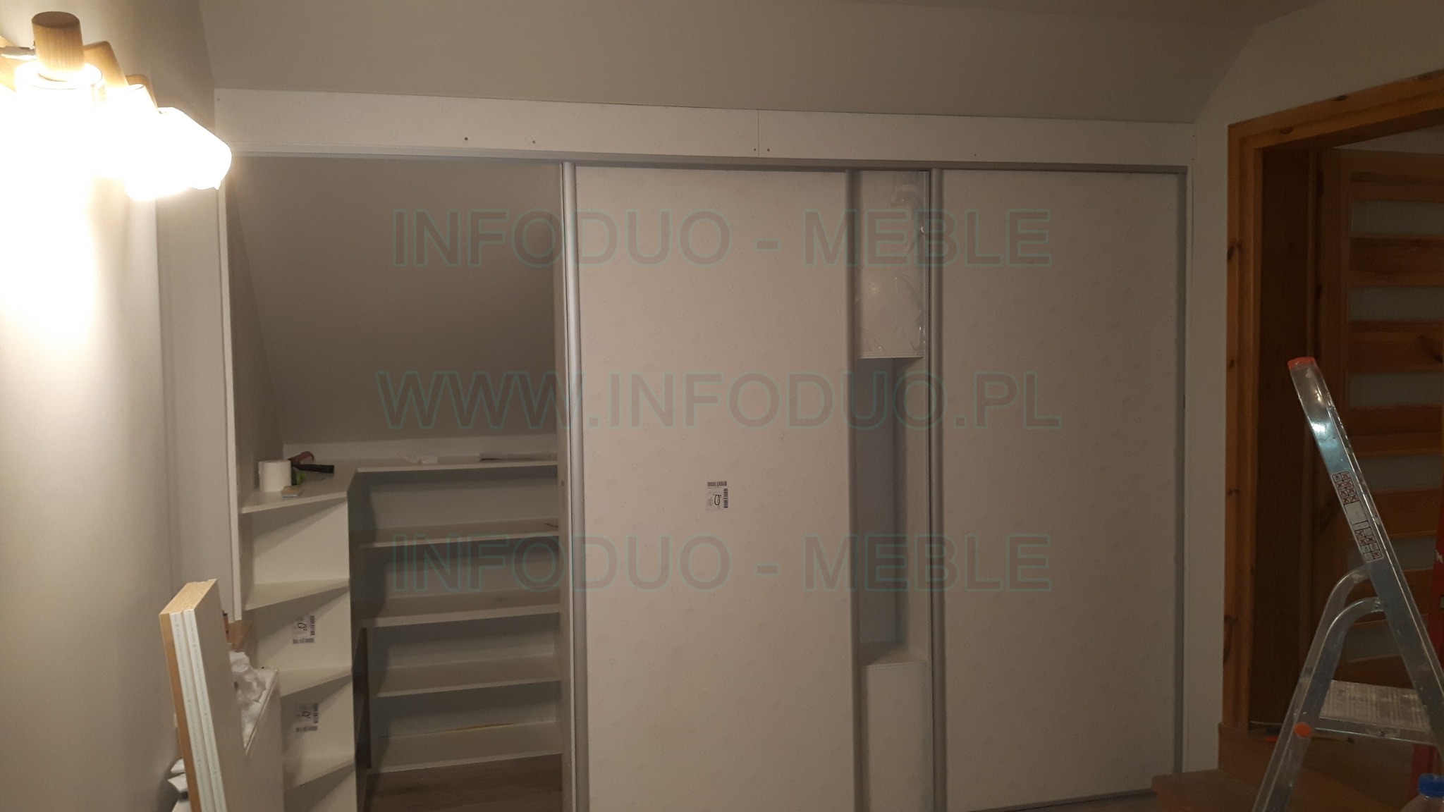Szafa przesuwna z białymi drzwiami w skosie poddasza, widoczne półki wewnątrz, drabina oparta o ścianę, montaż w trakcie.