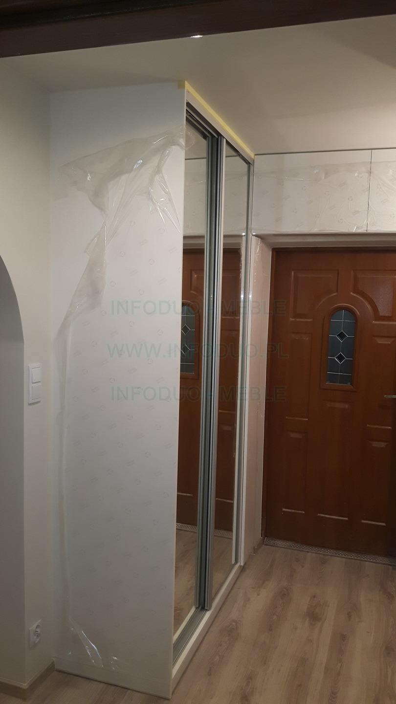 Nowa biała szafa z przesuwnymi lustrzanymi drzwiami, częściowo zabezpieczona folią ochronną, zamontowana we wnęce obok brązowych drzwi wejściowych.