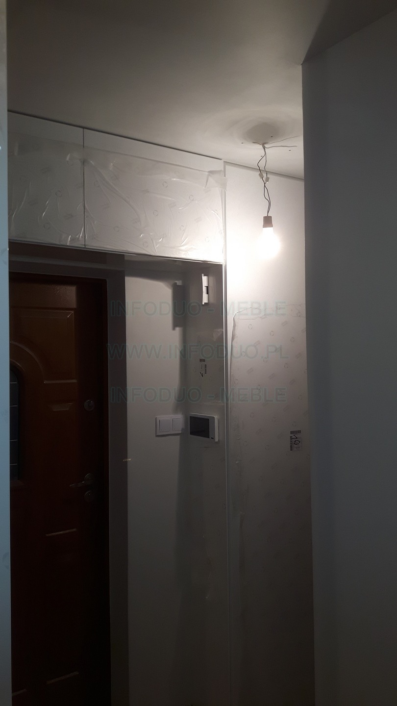 Montaż szafek pod sufitem w Lubartowie, zabezpieczonych folią ochronną, widoczny również fragment brązowych drzwi i surowa żarówka na kablu.