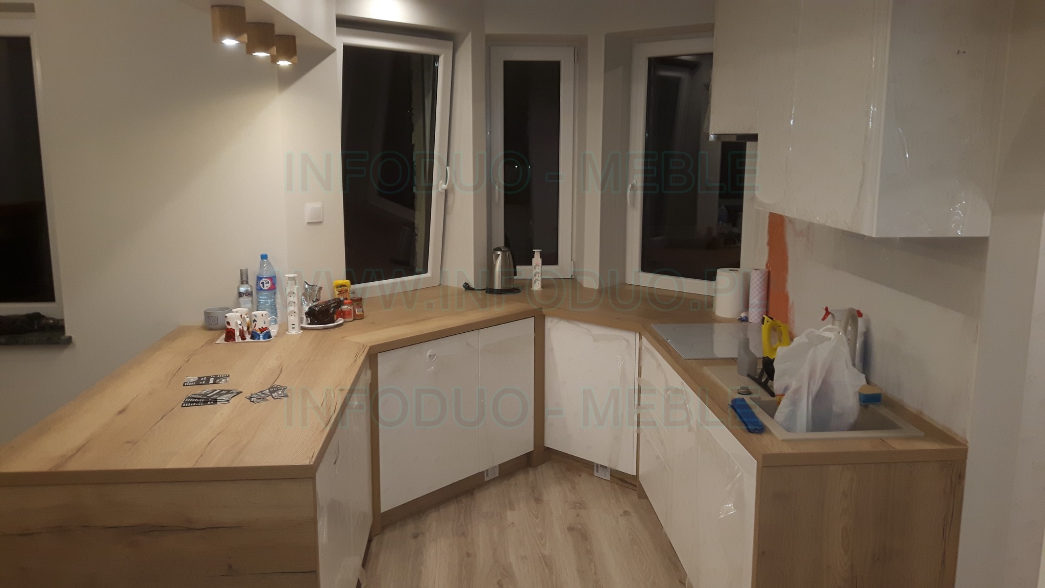 Nowoczesna kuchnia w trakcie montażu: białe szafki z drewnianymi blatami, folia ochronna na frontach, widoczne akcesoria kuchenne i okna w tle.