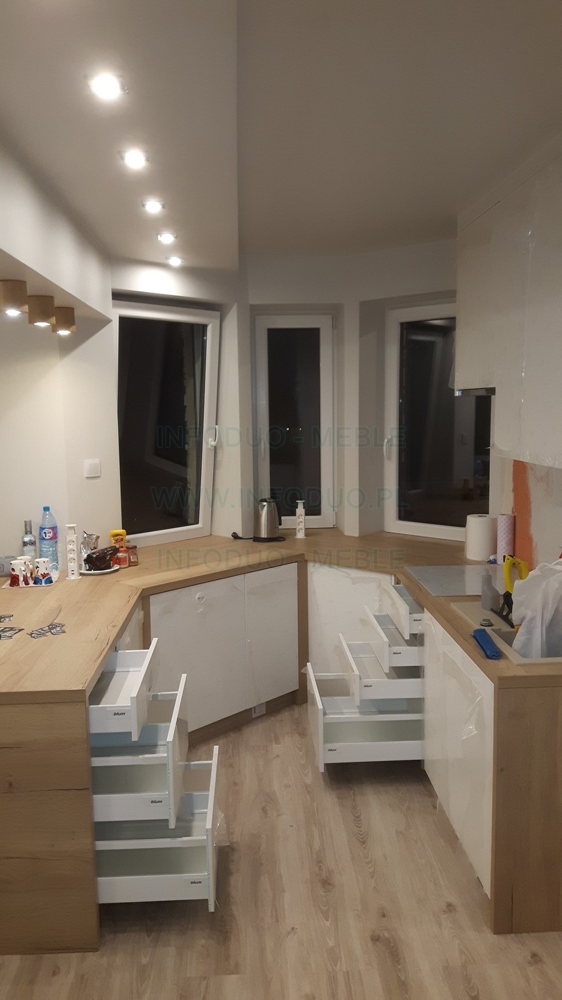 Jasna kuchnia z drewnianym blatem i białymi frontami, otwarte szuflady z systemem Blum, widoczne okna i akcesoria kuchenne na blacie.