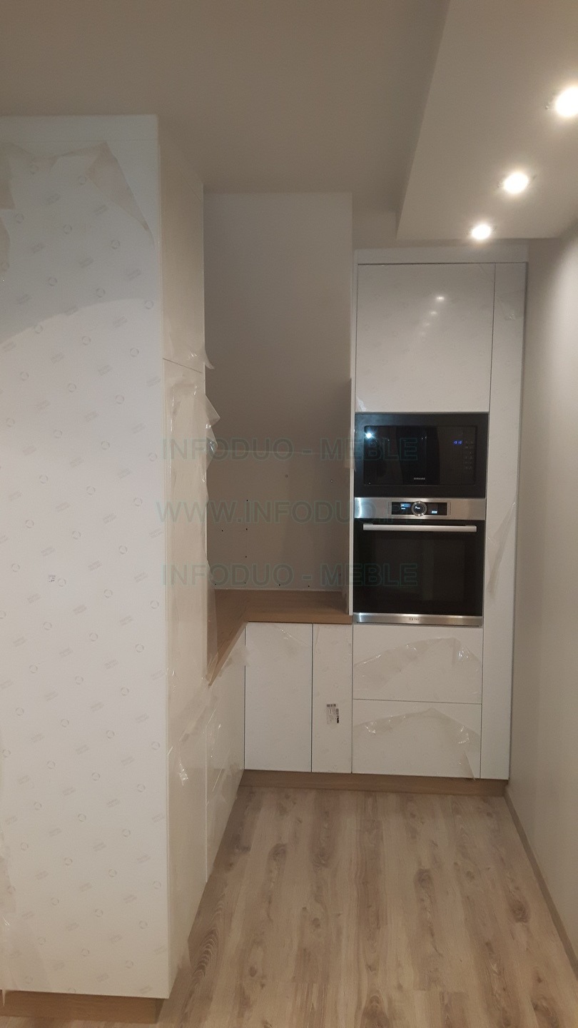 Biała zabudowa kuchenna z piekarnikiem i mikrofalówką w trakcie montażu, zabezpieczona folią ochronną, widoczna część drewnianego blatu.