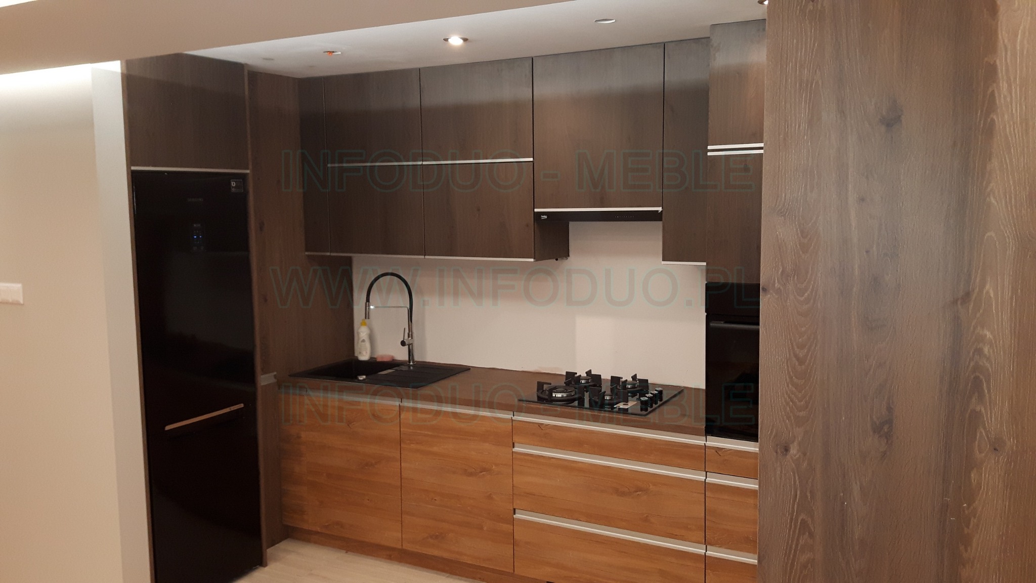 Nowoczesna kuchnia z drewnianymi frontami w dwóch odcieniach brązu, czarnym zlewem i sprzętem AGD, oświetlona punktowymi lampami sufitowymi.