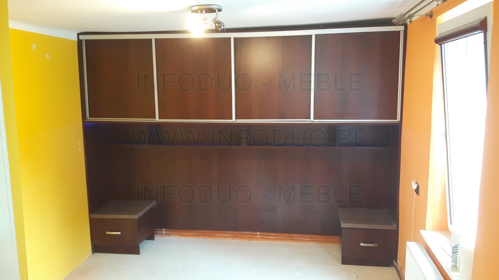 Zabudowa ściany z szafkami przesuwnymi i szafkami nocnymi w kolorze ciemnego drewna, na ścianie żółta farba, widoczne niedoskonałości wykończenia, na zdjęciu widoczny znak wodny.
