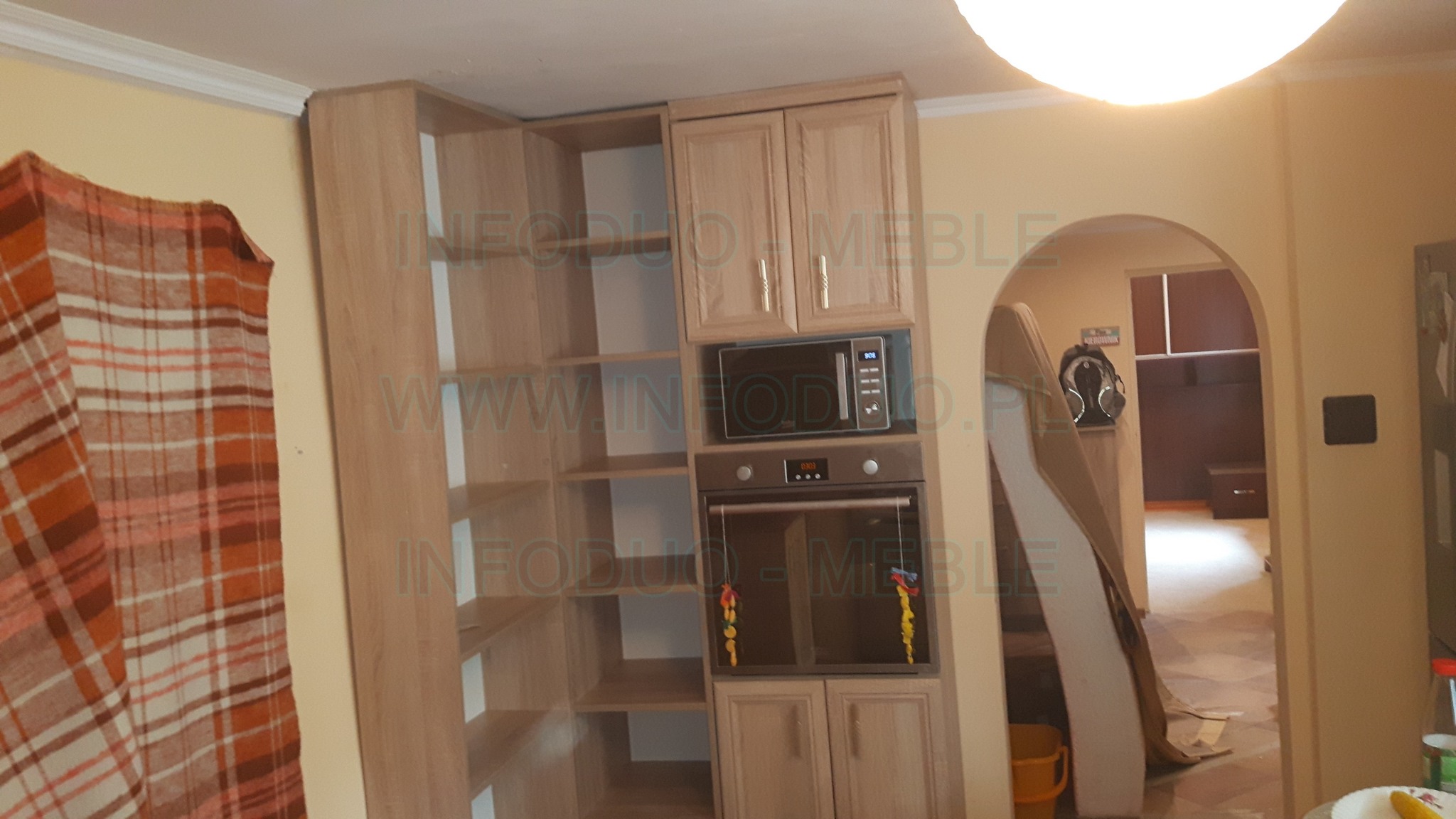 Jasna zabudowa kuchenna z piekarnikiem i mikrofalówką, po lewej stronie regał z półkami, widok na wnętrze przez łukowate przejście.