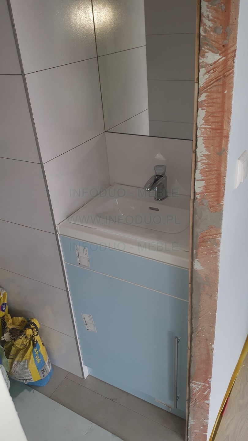 Łazienkowa szafka pod umywalkę w odcieniach błękitu, z widoczną armaturą i torbami z zaprawą w tle, w trakcie montażu, z surowym wykończeniem framugi.