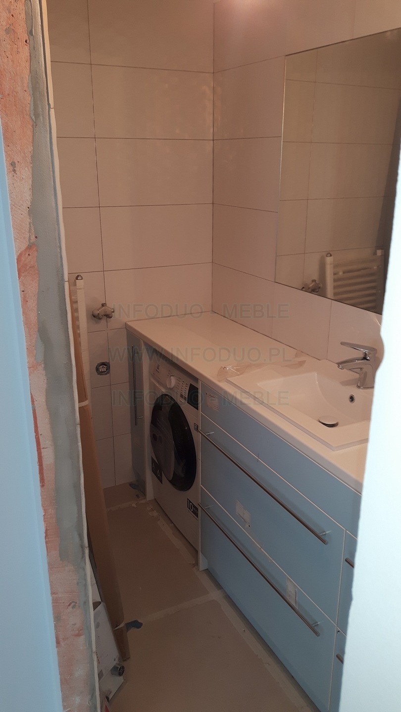 Łazienka w trakcie remontu: pralka zabudowana pod blatem z umywalką, niebieskie szafki z poziomymi uchwytami, widoczne niedokończone wykończenia ścian.