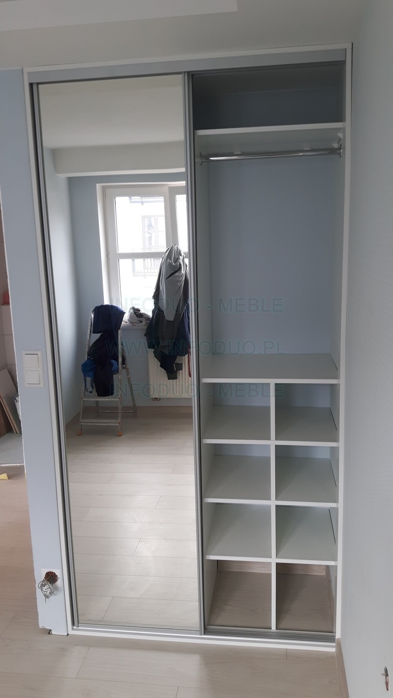 Biała szafa wnękowa z przesuwnymi drzwiami z lustrem, widoczna pusta przestrzeń z półkami i drążkiem, w lustrze odbicie pokoju z drabiną i ubraniami.