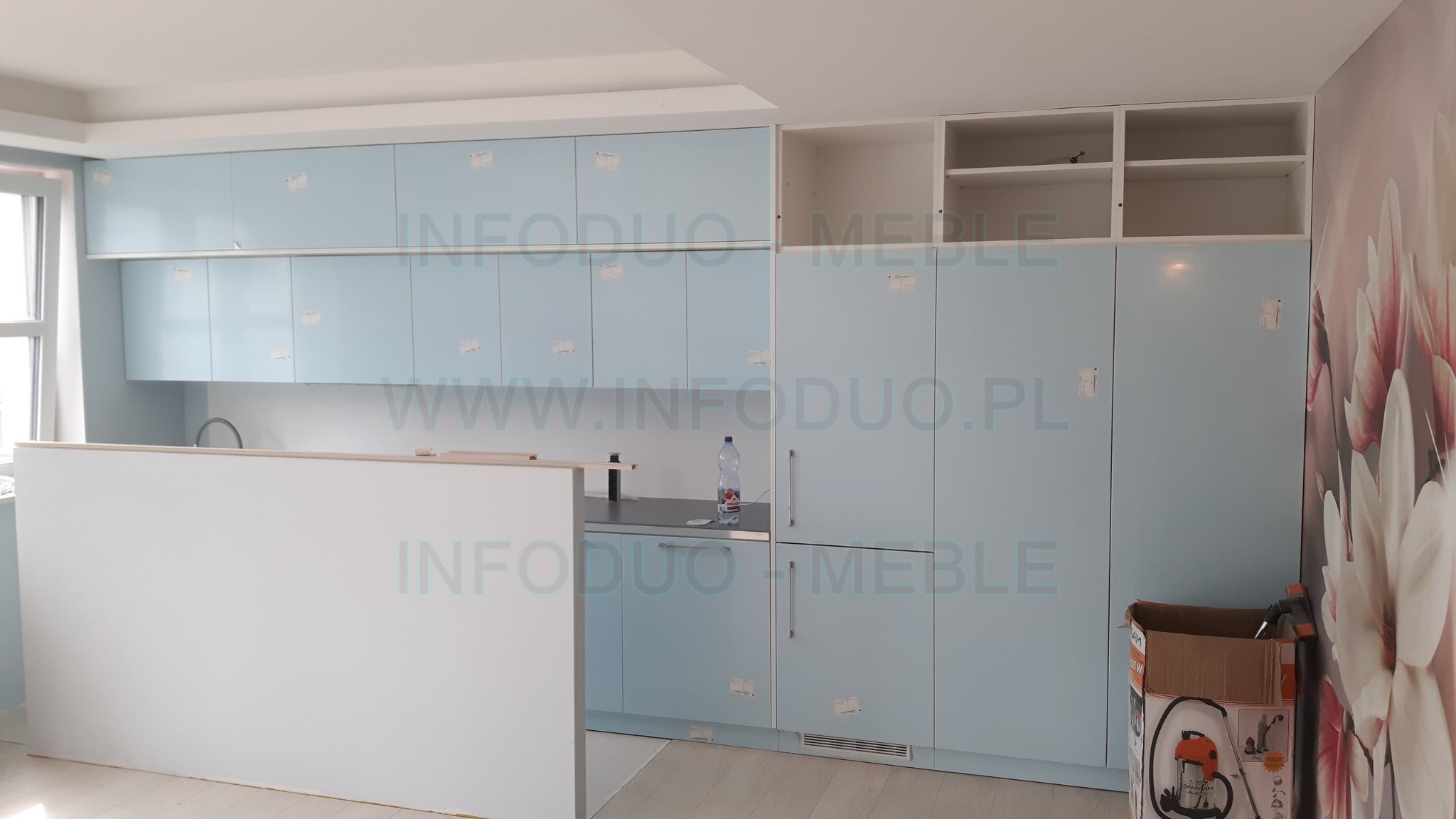 Niedokończona zabudowa kuchenna z błękitnymi frontami, widoczne kartony i pozostałości po montażu, ściana z fototapetą w kwiaty.