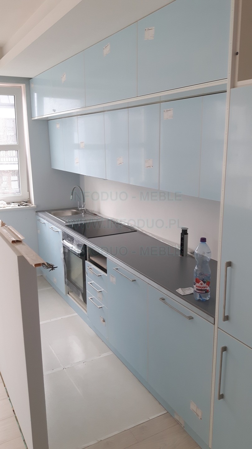 Niebieska zabudowa kuchenna z szarym blatem, widoczny zlew, płyta indukcyjna i piekarnik w trakcie montażu, podłoga zabezpieczona folią, butelka wody na blacie.