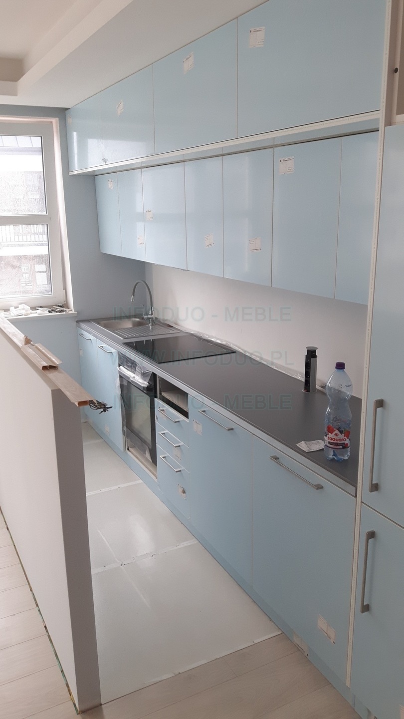 Jasnoniebieska zabudowa kuchenna z szarym blatem w trakcie montażu, widoczne niedokładności wykończenia i butelka wody na blacie.