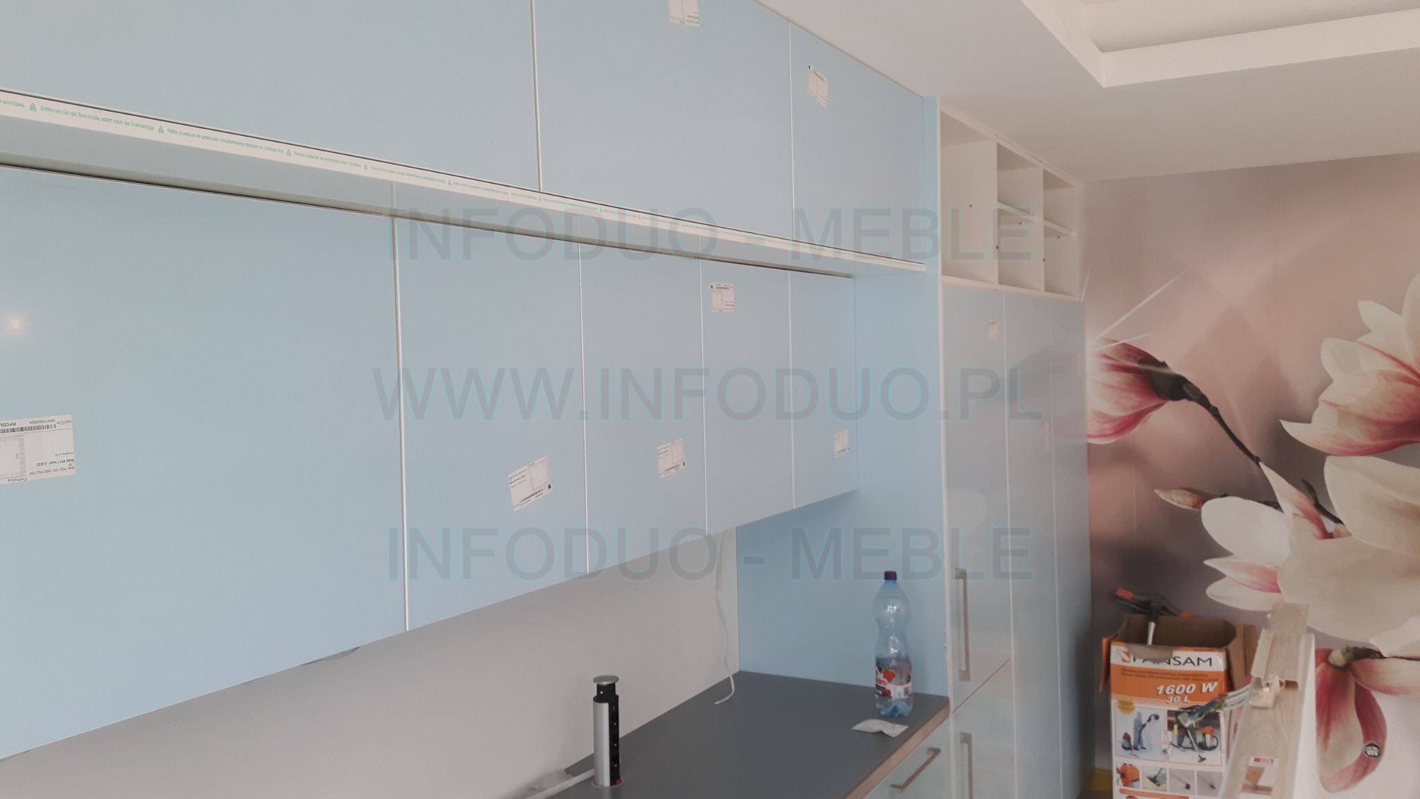 Jasnoniebieskie, lakierowane szafki kuchenne wiszące, częściowo zamontowane, z widocznymi etykietami ochronnymi, na tle ściany z tapetą w kwiaty magnolii i kartonem po odkurzaczu.