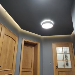 Kobar - Wnętrze korytarza z trzema drewnianymi drzwiami, szarą ścianą i ciemnym sufitem z okrągłą lampą oraz oświetleniem LED wzdłuż krawędzi.