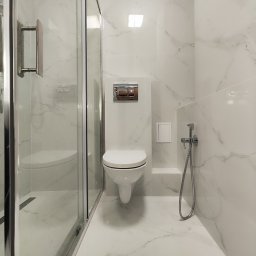 Kobar - Nowoczesna łazienka z białymi płytkami imitującymi marmur, wisząca toaleta z chromowanym przyciskiem spłukiwania, szklana kabina prysznicowa z chromowanymi okuciami i bidetka.